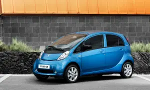 En blå Peugeot iOn står parkerad framför en grå tegelvägg med en orange takkant.