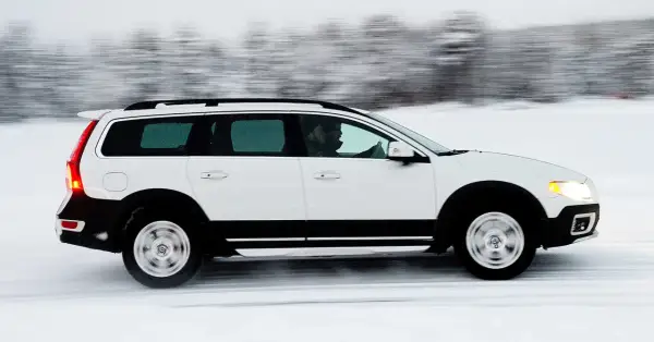 Vit Volvo XC70 kör på en snöig väg, med en förare bakom ratten. Bilen är i rörelse, vilket skapar en suddig bakgrund av träd.